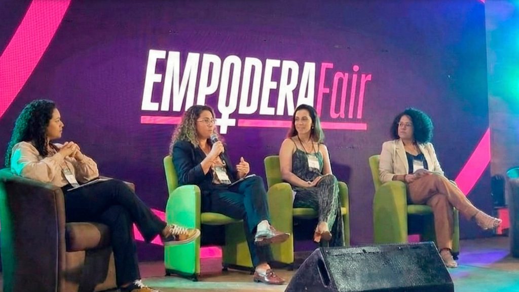 Quatro mulheres sentadas em poltronas no palco da Empodera Fair, com o logotipo do evento ao fundo, durante painel de debate