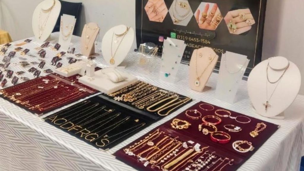 Estande com colares, pulseiras e anéis expostos em displays sobre mesa na área de exposição
