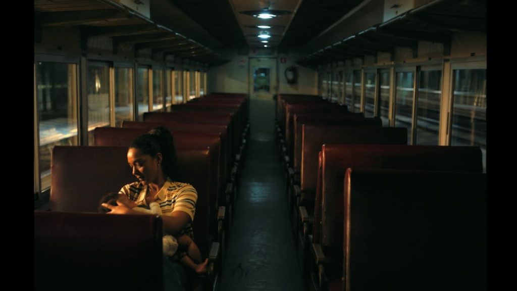 Mulher sentada em um vagão de trem vazio e pouco iluminado, segurando um bebê no colo de forma afetuosa. Cena do filme Despedidas da campanha de Dia das Mães do Boticário.