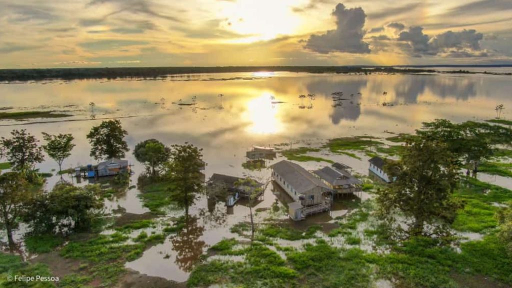 Vista aérea de comunidade ribeirinha na Ilha das Guaribas, Rio Amazonas, com casas sobre palafitas cercadas pela cheia, vegetação verde e pôr do sol refletido nas águas