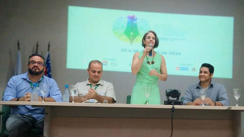 Mesa de debates em um auditório com quatro pessoas. Uma mulher de vestido verde está de pé falando ao microfone, enquanto os outros três homens, sentados, prestam atenção. Ao fundo, um telão projeta a logomarca de um evento acadêmico.