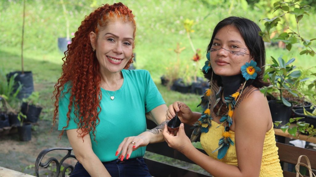 A jornalista Edilene Mafra sorri enquanto a jovem indígena Tainara Kambeba aplica uma pintura tradicional em seu braço. Ambas estão sentadas ao ar livre, cercadas por plantas.