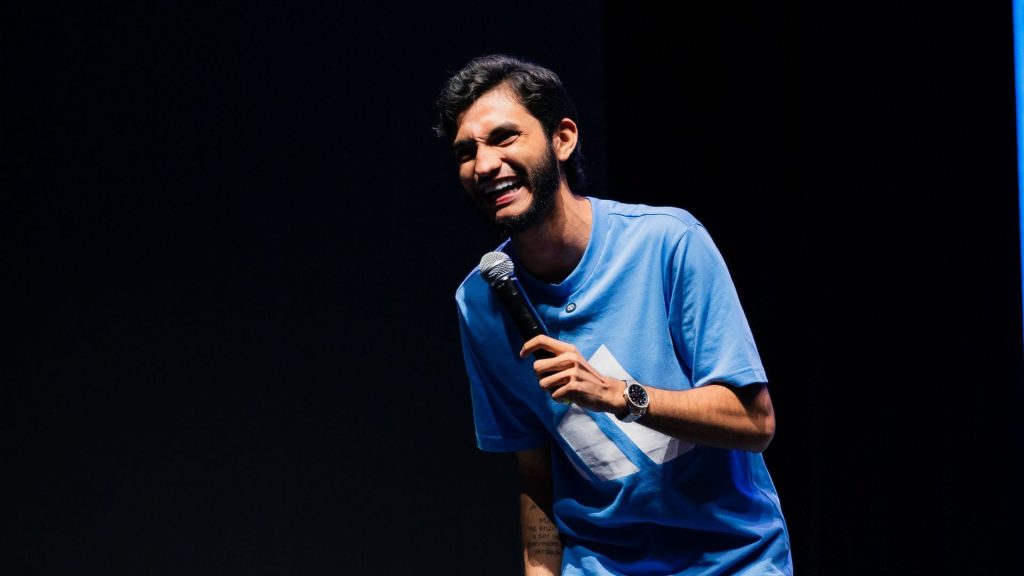 O humorista amazonense Leandro Leitte sorrindo muito com um microfone na mão durante apresentação de stand-up comedy.