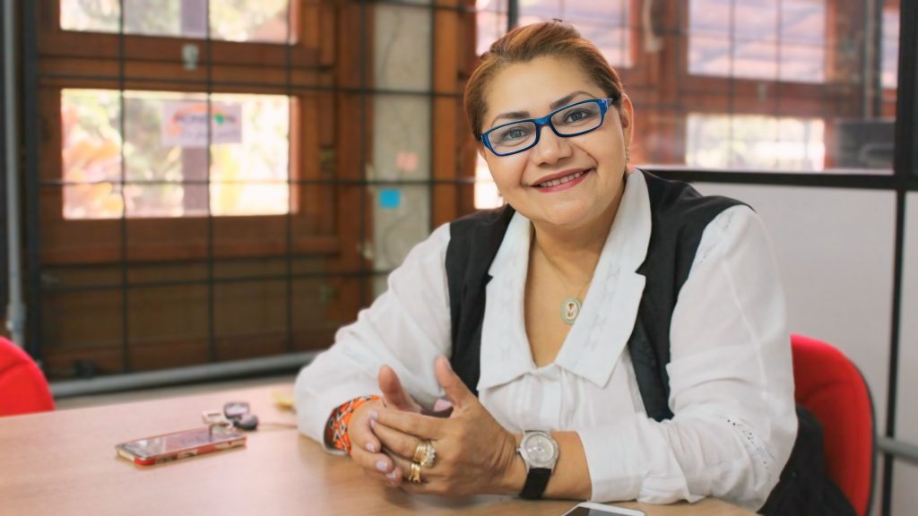 Marilene Corrêa da Silva Freitas, professora da UFAM e Pesquisadora Emérita do CNPq, sentada a uma mesa durante entrevista, sorrindo e gesticulando com as mãos, usando blusa branca, colete preto e óculos azuis