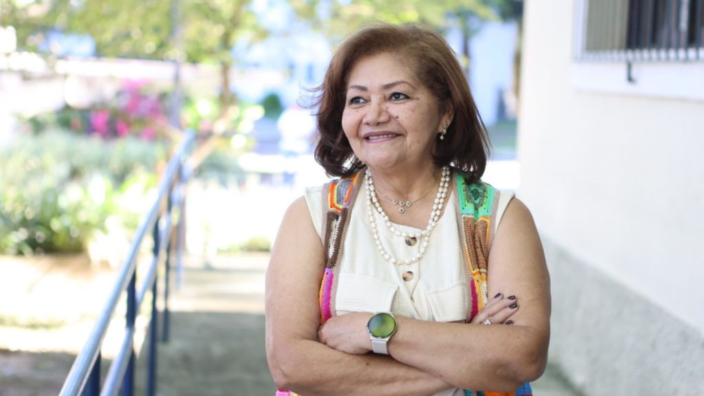Marilene Corrêa da Silva Freitas, pesquisadora da UFAM, de braços cruzados e sorrindo, usando colete colorido artesanal e colar de pérolas, em área externa com vegetação ao fundo