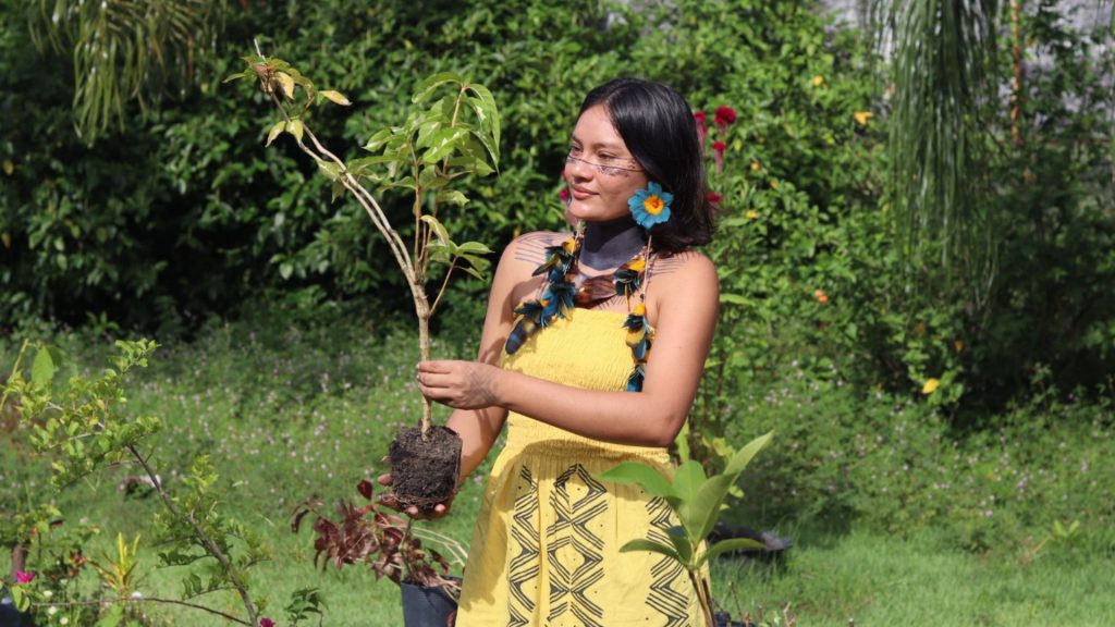 A jovem ativista indígena Tainara Kambeba segura cuidadosamente uma muda de planta com raiz em um jardim ensolarado, simbolizando seu compromisso com a sustentabilidade e a preservação ambiental.