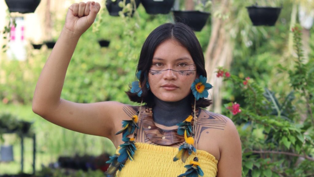 Tainara Kambeba, jovem indígena com trajes e pinturas tradicionais, ergue o punho direito cerrado em um gesto de força, resistência e luta por direitos, tendo a natureza como pano de fundo.