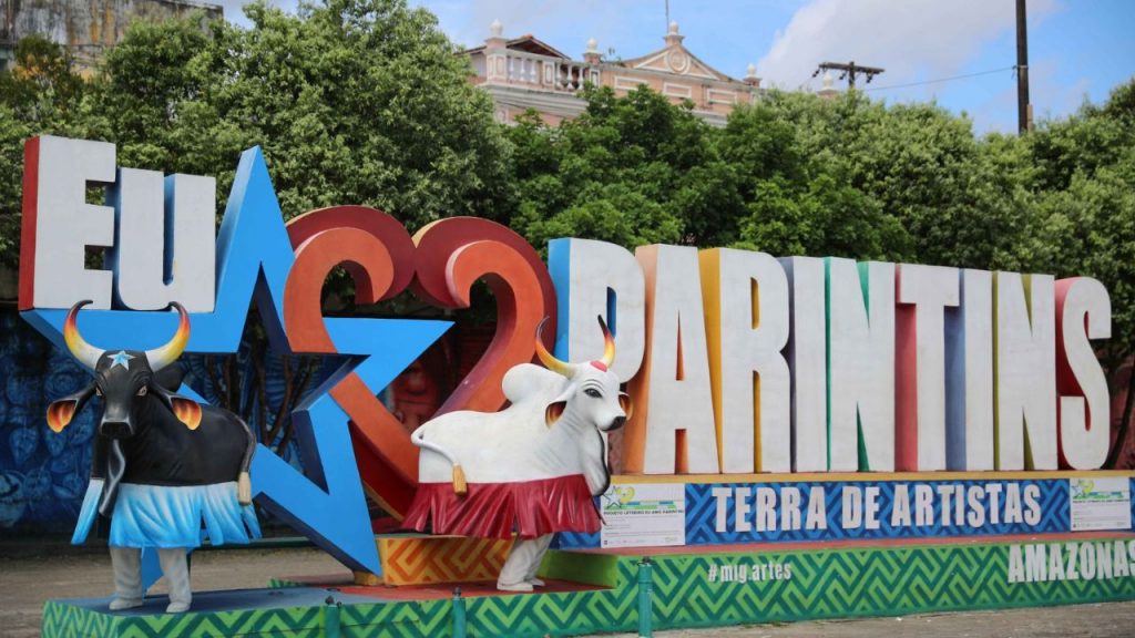 Letreiro colorido de Parintins com as letras em cores vibrantes, apresentando as figuras dos bois Caprichoso em azul e Garantido em branco, símbolos principais do Festival de Parintins, com a inscrição 'Terra de Artistas' e 'Amazonas' na base