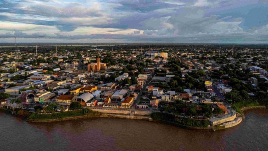 Fotografia aérea da Orla de Parintins na Ilha Tupinambarana, mostrando a cidade com suas estruturas coloridas, vegetação amazônica densa, Rio Amazonas em primeiro plano, e céu nublado típico da região, demonstrando o território e identidade amazônica