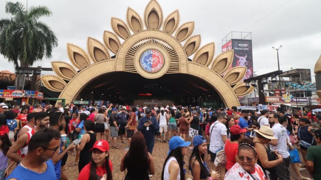 Multidão de turistas e visitantes na Praça da Matriz de Parintins durante o Festival, com estrutura de palco em forma de flor dourada, pessoas usando cores do Boi Caprichoso (azul) e Boi Garantido (vermelho), demonstrando a vivência autêntica e emoção coletiva do evento
