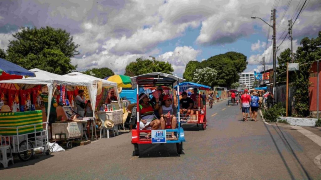 Transporte típico de Parintins - tuc-tuc em cores azul e vermelho - em rua movimentada durante o Festival, com visitantes, barracas comerciais, estruturas de tendas coloridas, demonstrando a vivência autêntica e o conhecimento local que a OCA Lazer proporciona